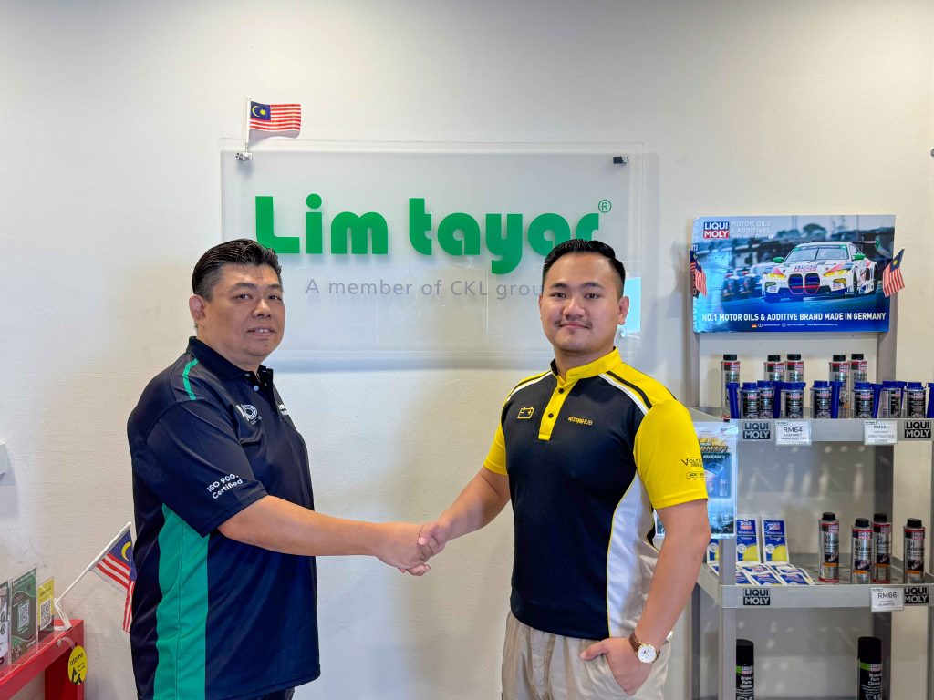 Two representatives from Lim Tayar and Baterihub Xpress are shaking hands in front of a Lim Tayar sign, symbolizing their partnership. The Lim Tayar representative wears a black and green polo shirt, while the Baterihub Xpress representative wears a black and yellow uniform. A shelf displaying automotive products, including Liqui Moly motor oils and additives, is visible in the background, along with small Malaysian flags, reinforcing the local branding.