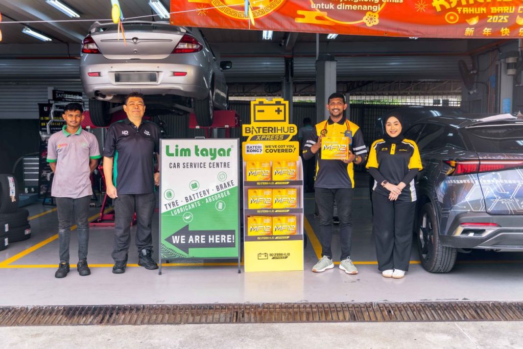 A group of four people stands in front of a car service center named "Lim Tayar." Two of them, wearing black and yellow uniforms, represent Baterihub Xpress, while the other two, in black and grey-green uniforms, are from Lim Tayar. The setting includes a car lifted on a hydraulic lift and another car parked nearby. A promotional stand with Baterihub Xpress branding and car batteries is prominently displayed in the center. The group is smiling, with one person holding a battery product, indicating a collaboration between Baterihub and Lim Tayar. The background features festive Chinese New Year decorations.