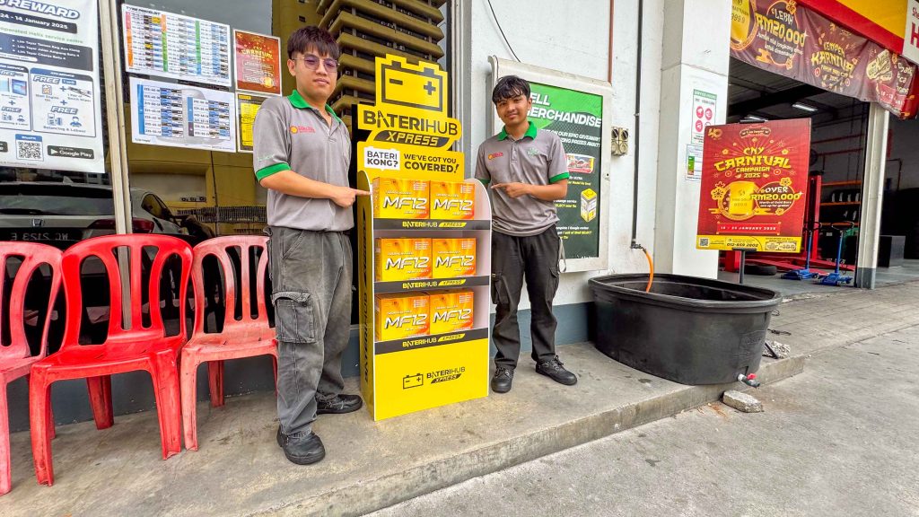 Two automotive service employees in grey and green uniforms stand outside a Lim Tayar service center, pointing at a Baterihub Xpress battery display. The display features MF12 car batteries with a bright yellow and black design.